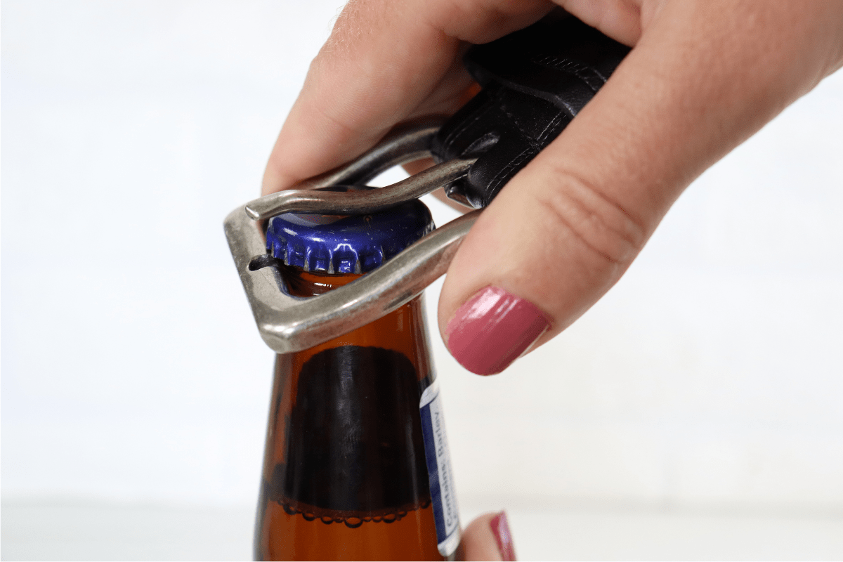woman hand with pink nail polish using belt buckle to open beer bottle white background