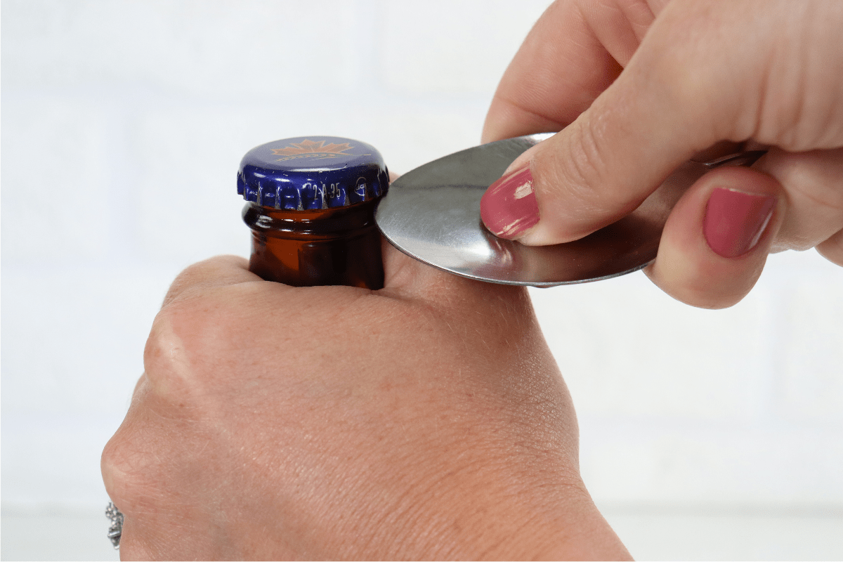 woman with pink nail polish opening beer bottle with metal spoon white background