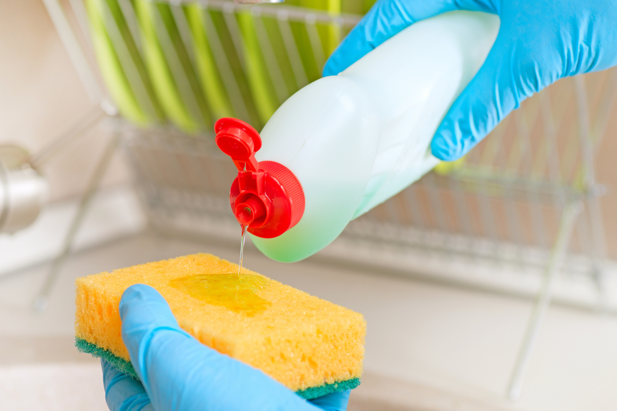 blue gloved hands pouring dish soap onto yellow sponge dish rack blurred in background