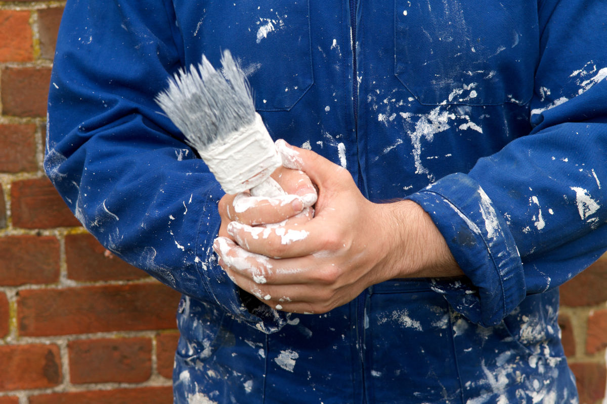 close up of man holding paint brush with white paint all over his coveralls