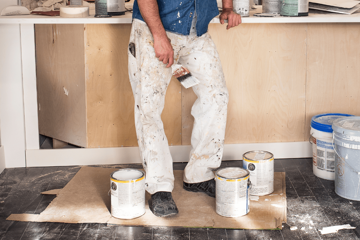 man's legs wearing white painting pants standing with brush in hand on brown kraft paper with cans of paint around messy floor