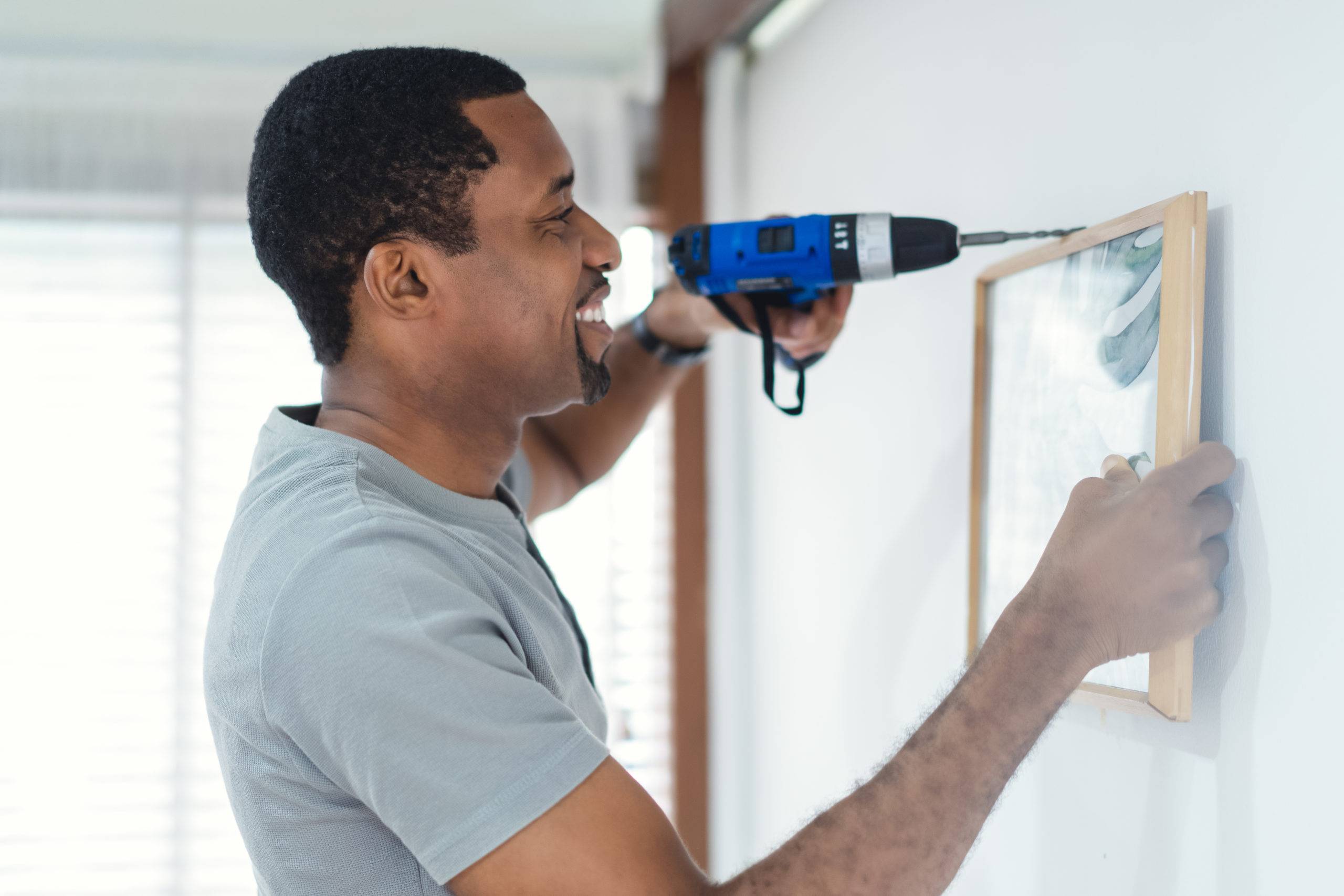 Smiling black man builder drilling a hole in white wall and putting picture frame at home. Home renovation and decoration. Adult making DIY workshop. Concept for home diy, self service.