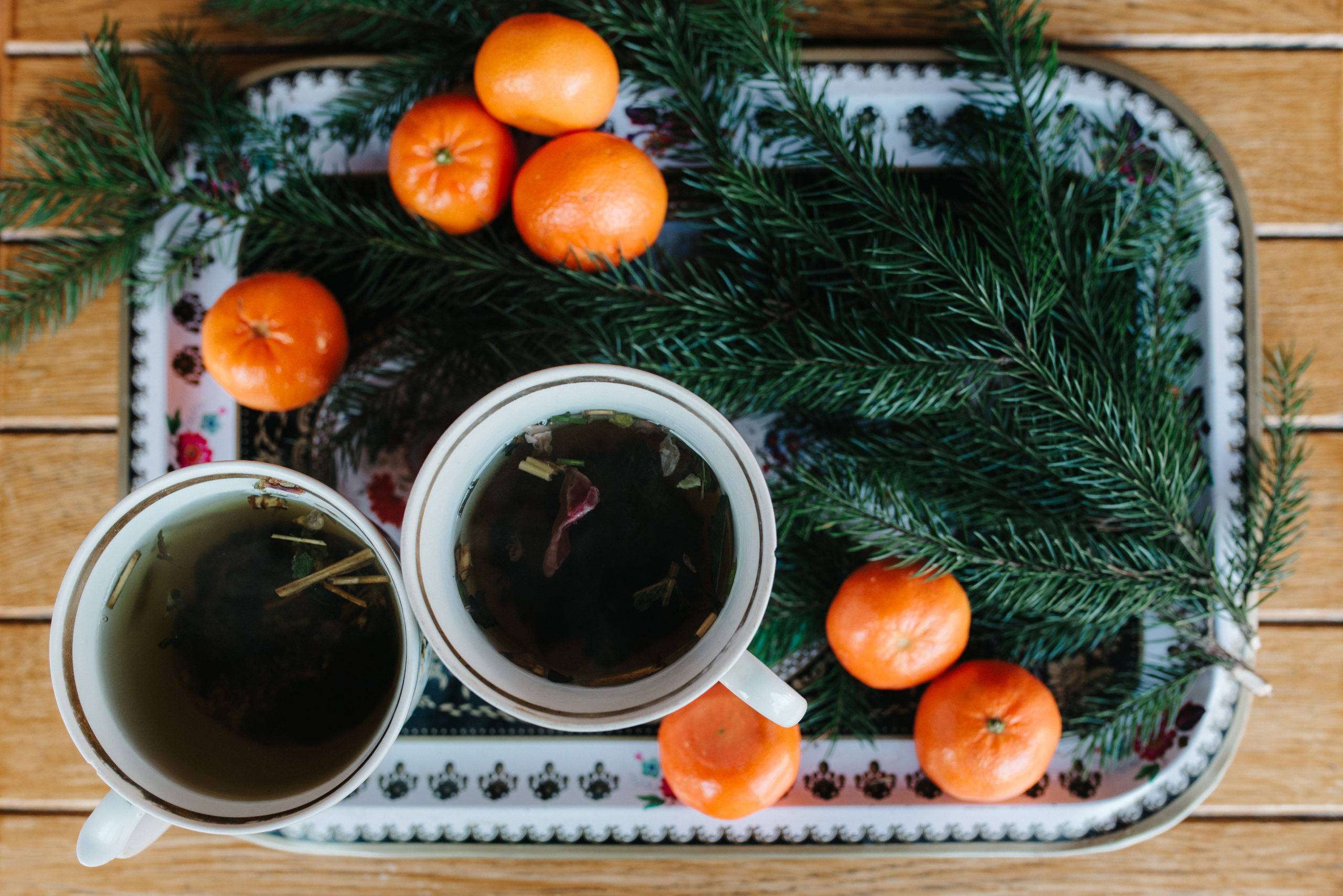 two cups of brewed tea on tray surrounded by tangerines and pine needle branches