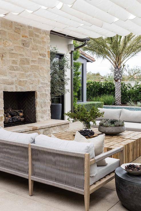low height outdoor sofa with white cushions, next to outdoor brick fireplace and white pergola overhead