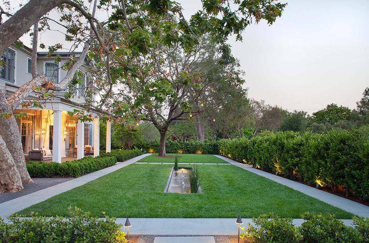 large backyard space with perfectly manicured green lawn, large tree set towards back of lawn, lined by potted lights
