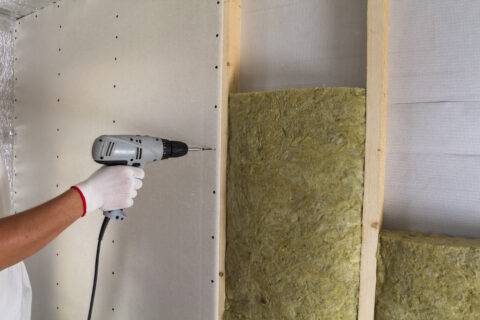 Close-up of worker hand with electric screwdriver fastening drywall to wooden timber frame with insulation mineral rock wool staff. Warm home, low energy bills, construction and renovation concept.