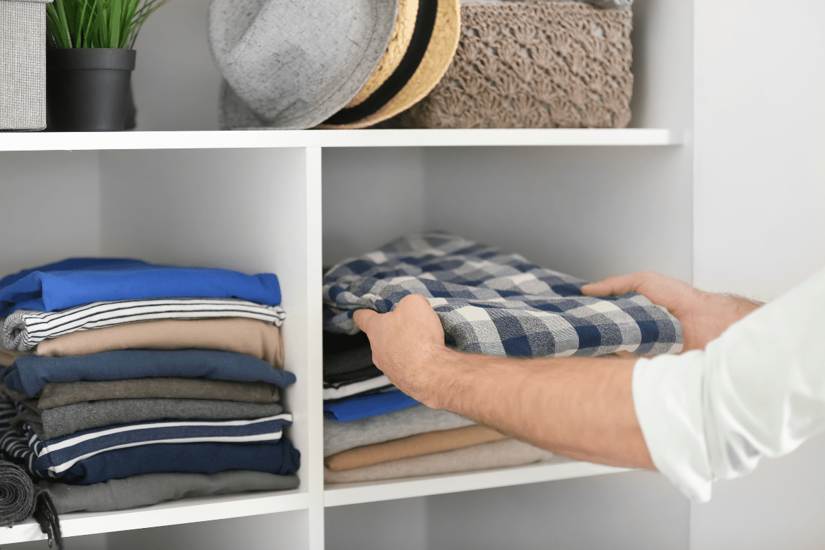 man placing folded shirt on top of stacked shirts in open white shelving 