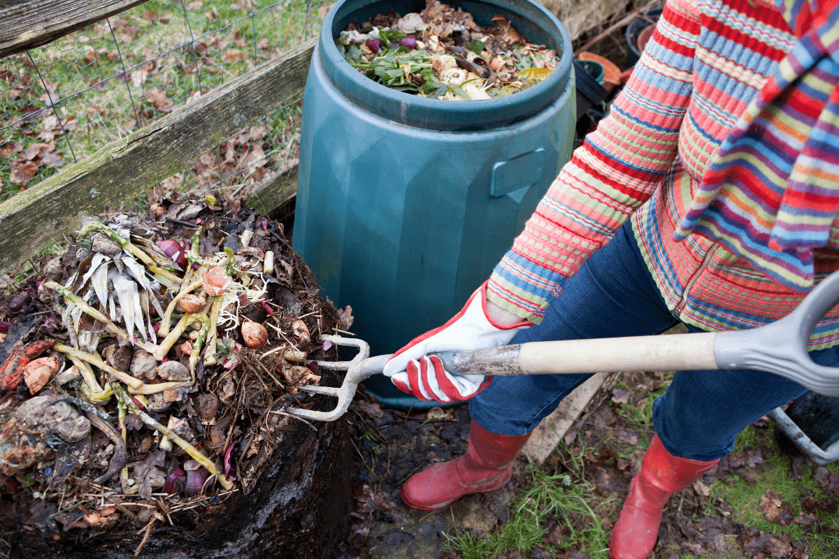 woman in brightly colored shirt with red rain boots and gardening gloves holding pitch fork, turning compost bin