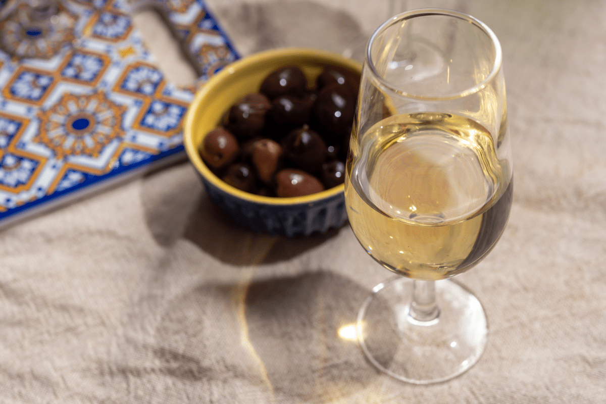 port glass of fortified wine, sitting on beige tablecloth next to ramekin of olives 