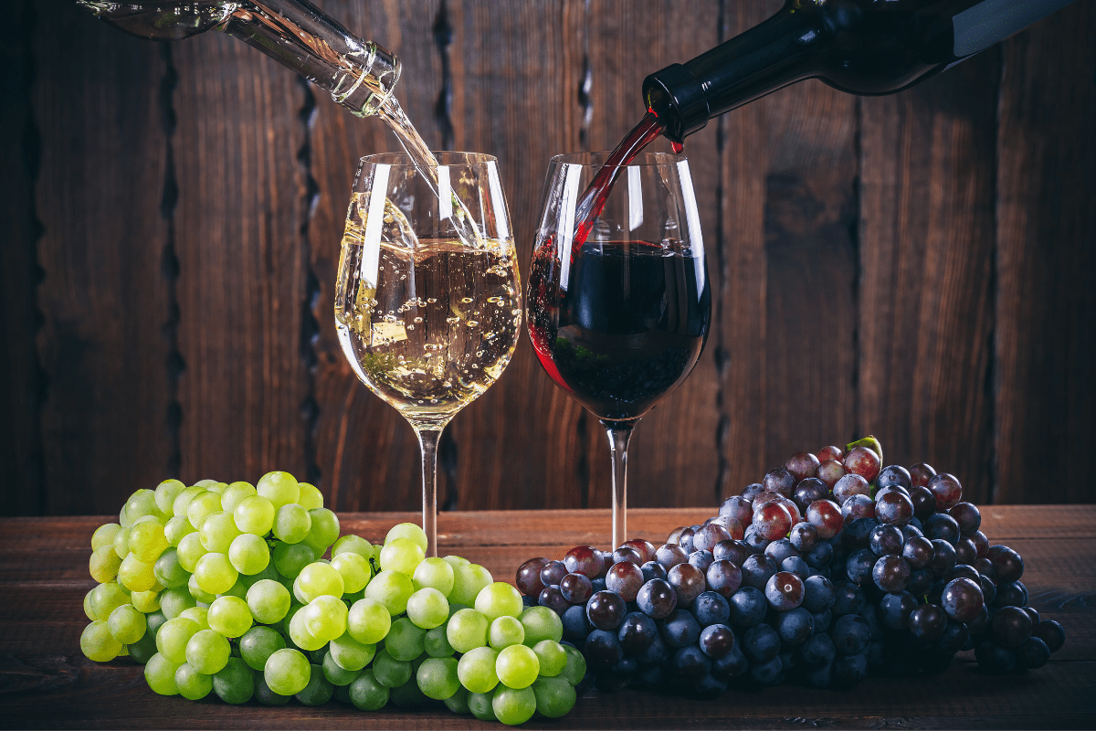 glass of white wine being poured next to glass of red wine, two bunches of grapes sitting at base of glasses