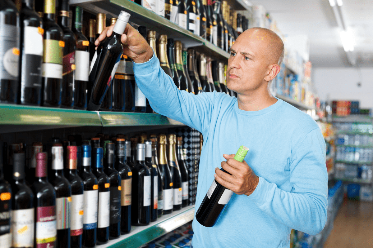 serious man in blue shirt choosing wine bottles from store aisle