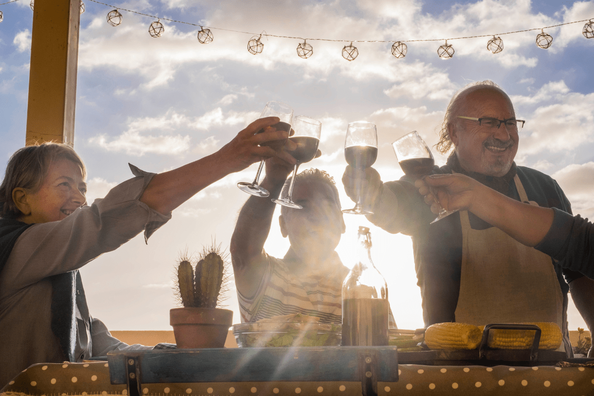 four older people on backyard patio holding up glasses of wine with sun shining in background