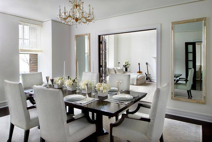 Elegant formal dining room with french doors to living room, flanked by a pair of tall champagne silver leaf wall mirrors, with a rectangular pedestal dining table in the center of the space lined with light gray arm dining chairs atop a tonal gray scroll patterned rug illuminated by a gold leafed crystal chandelier.