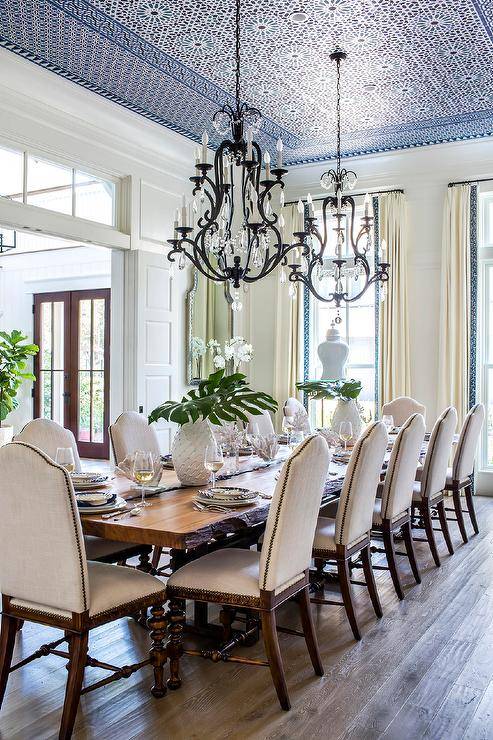 Two French crystal chandeliers hangs from a blue Moroccan style ceiling over a long live edge dining table surrounded by French camelback dining chairs, while windows are covered in ivory curtains.