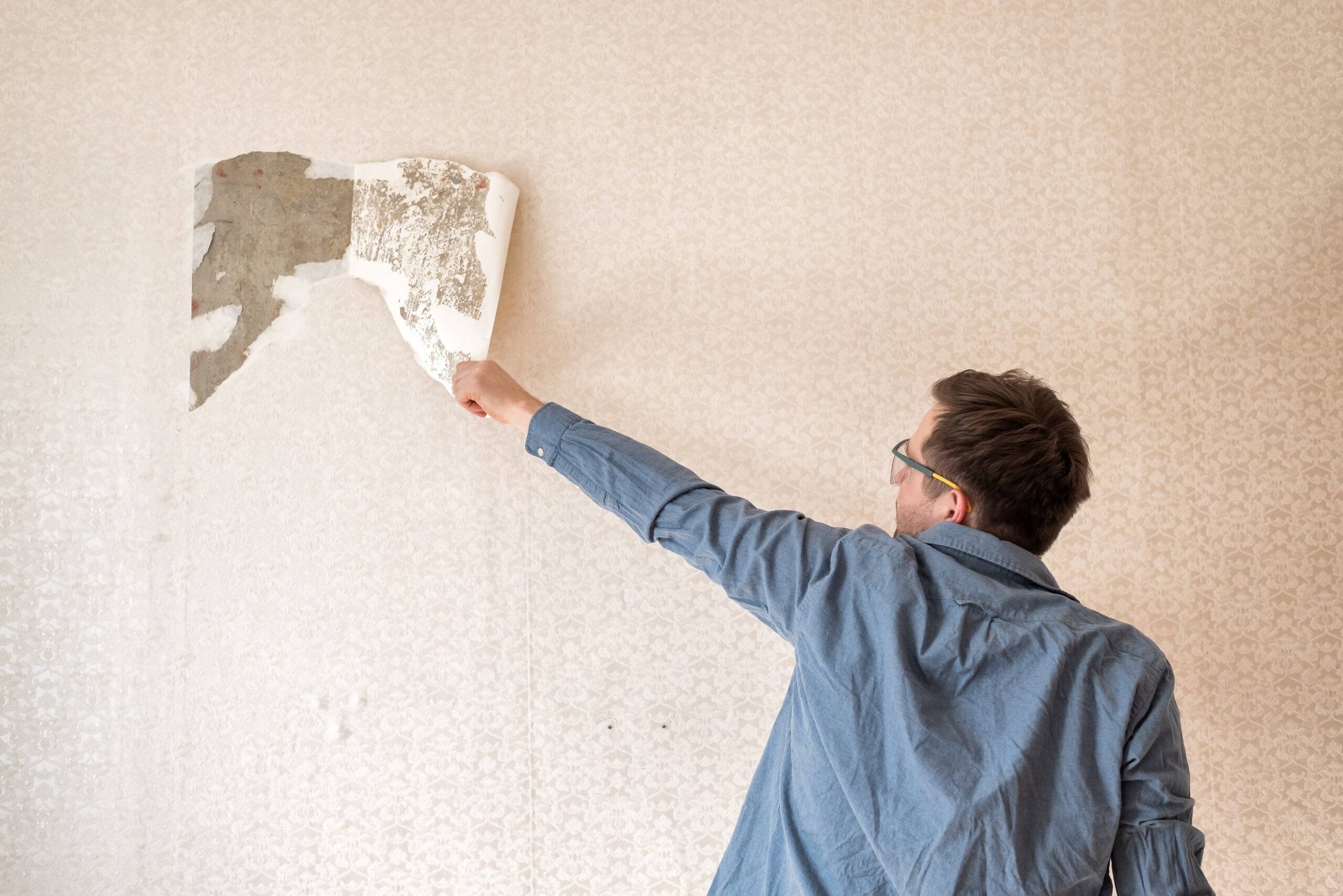 Process of flat renovation. Young man wearing safety glasses is tearing off old wallpaper from the wall preparing for home redecoration