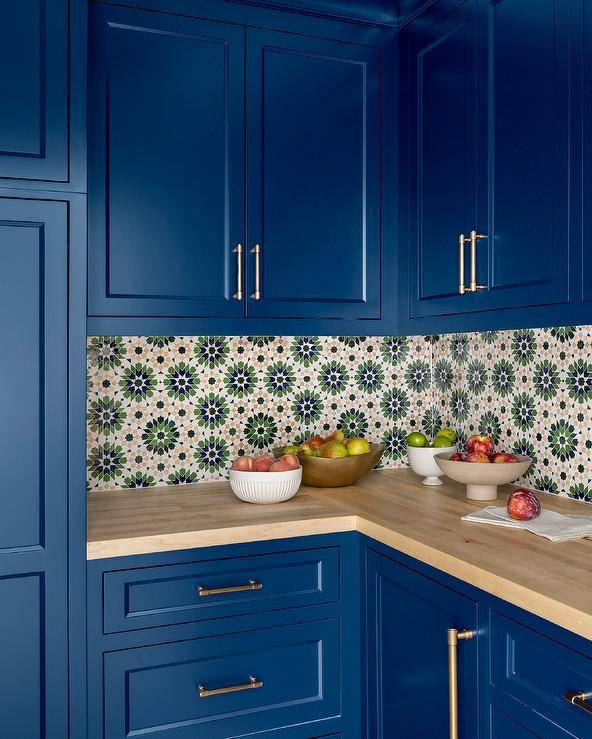 Bright blue kitchen cabinets are complemented with brass hardware and a maple butcher block countertop fixed against green and orange mosaic backsplash tiles under blue upper cabinets.