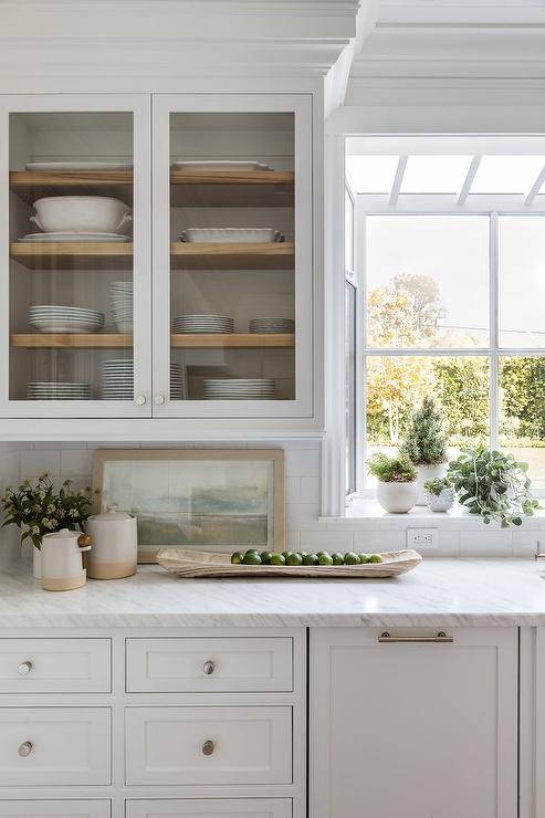 Glass front kitchen cabinets are mounted beside a garden window and over white cabinetry donning polished nickel hardware and a marble countertop.