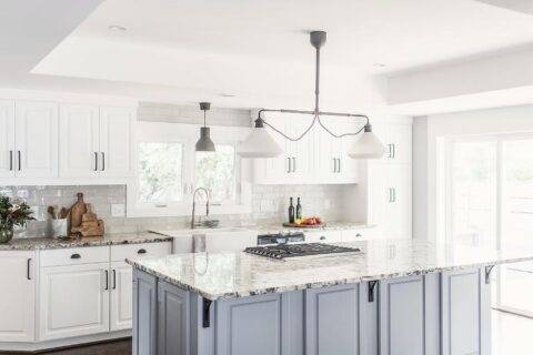 Kitchen features a blue kitchen island topped with white, gray and black granite with iron island corbels, kitchen island cooktop, illuminated by a linear schoolhouse lantern. The design is completed with white kitchen cabinets with black pulls, white apron sink and white gray and black granite countertop.