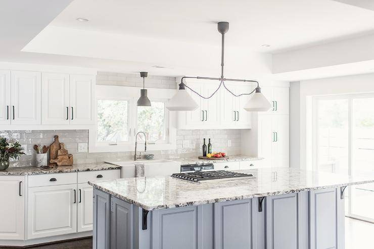 Kitchen features a blue kitchen island topped with white, gray and black granite with iron island corbels, kitchen island cooktop, illuminated by a linear schoolhouse lantern. The design is completed with white kitchen cabinets with black pulls, white apron sink and white gray and black granite countertop.