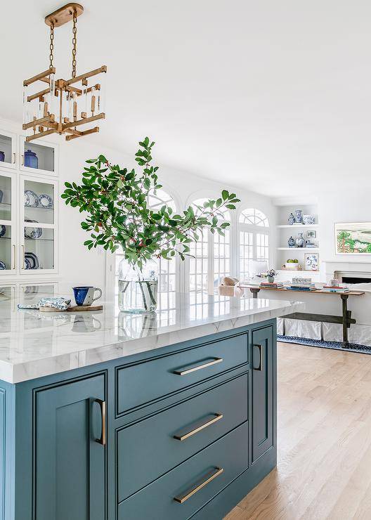 A blue kitchen island adorned with long brass pulls and a marble countertop is lit by a brass and lucite chandelier.