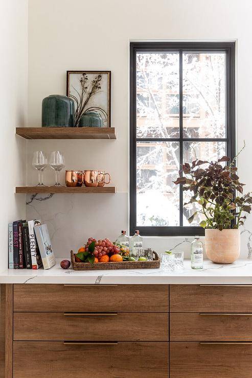 Kitchen features brown veneer cabinetry with marble look quartz countertops under a window and small floating shelves.