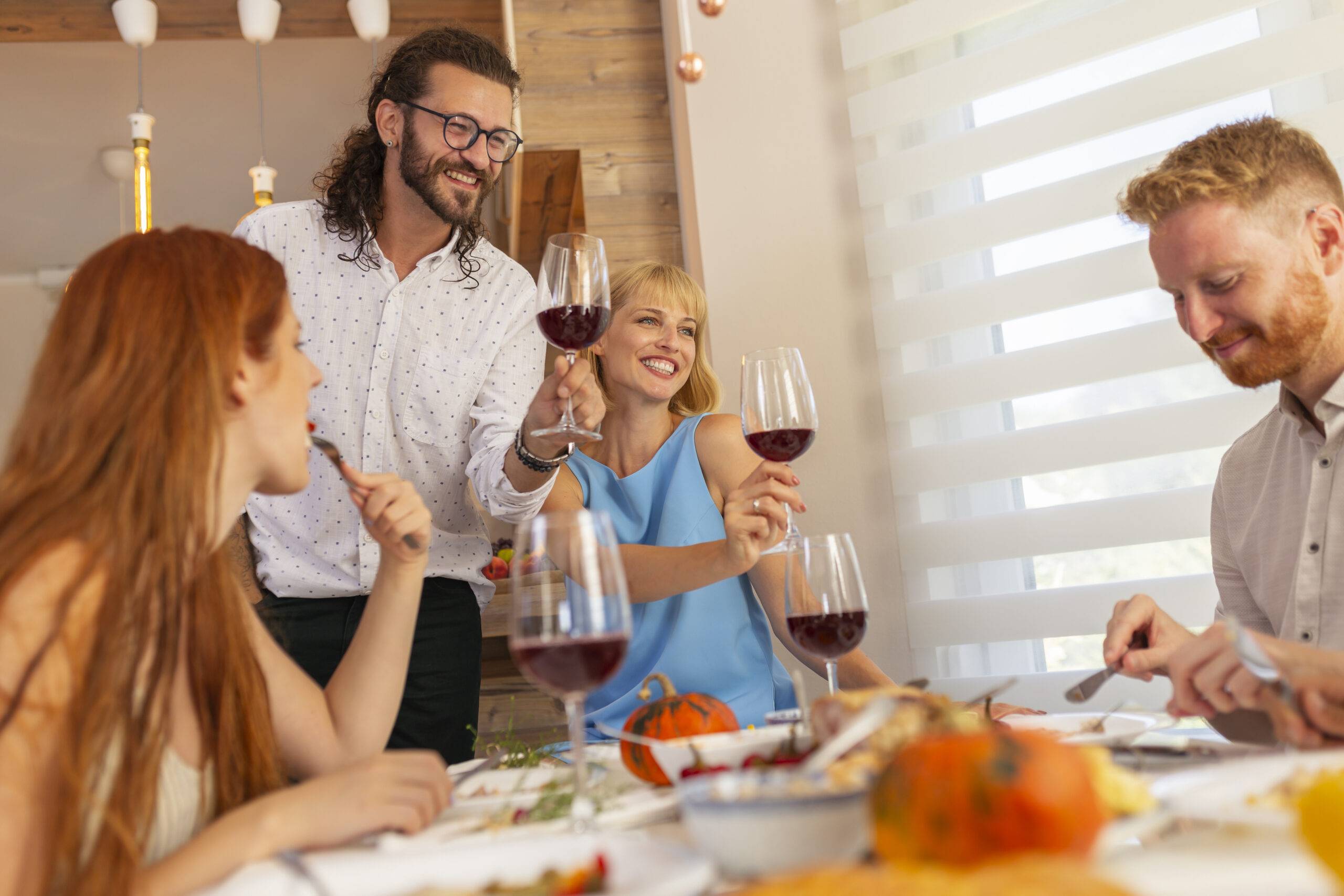 Group of young friends gathered around the table for Thanksgiving, hosts making a toast, raising glasses of wine and entertaining their guests.
