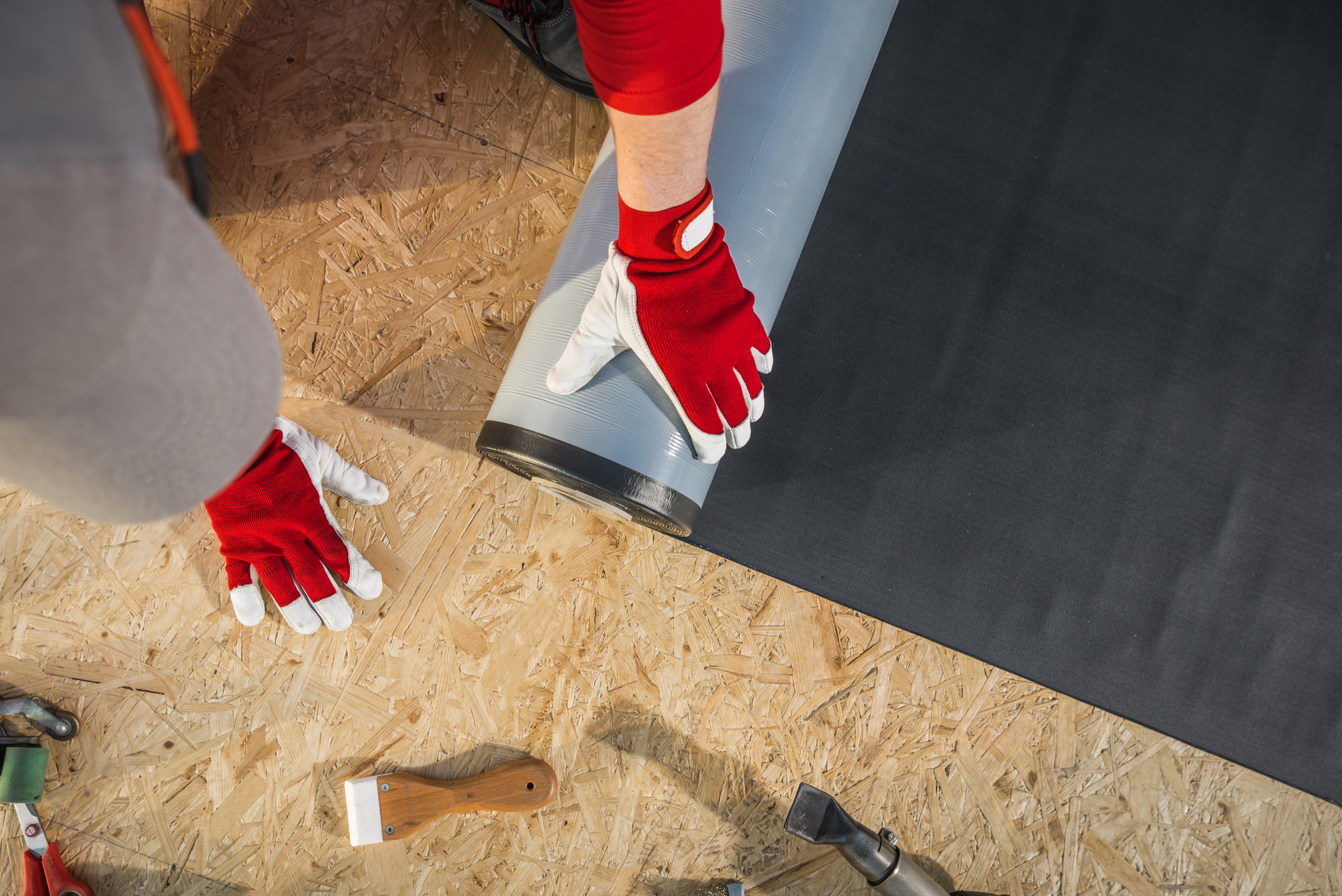 A person with gloves laying down vinyl roof layer on top of plywood.