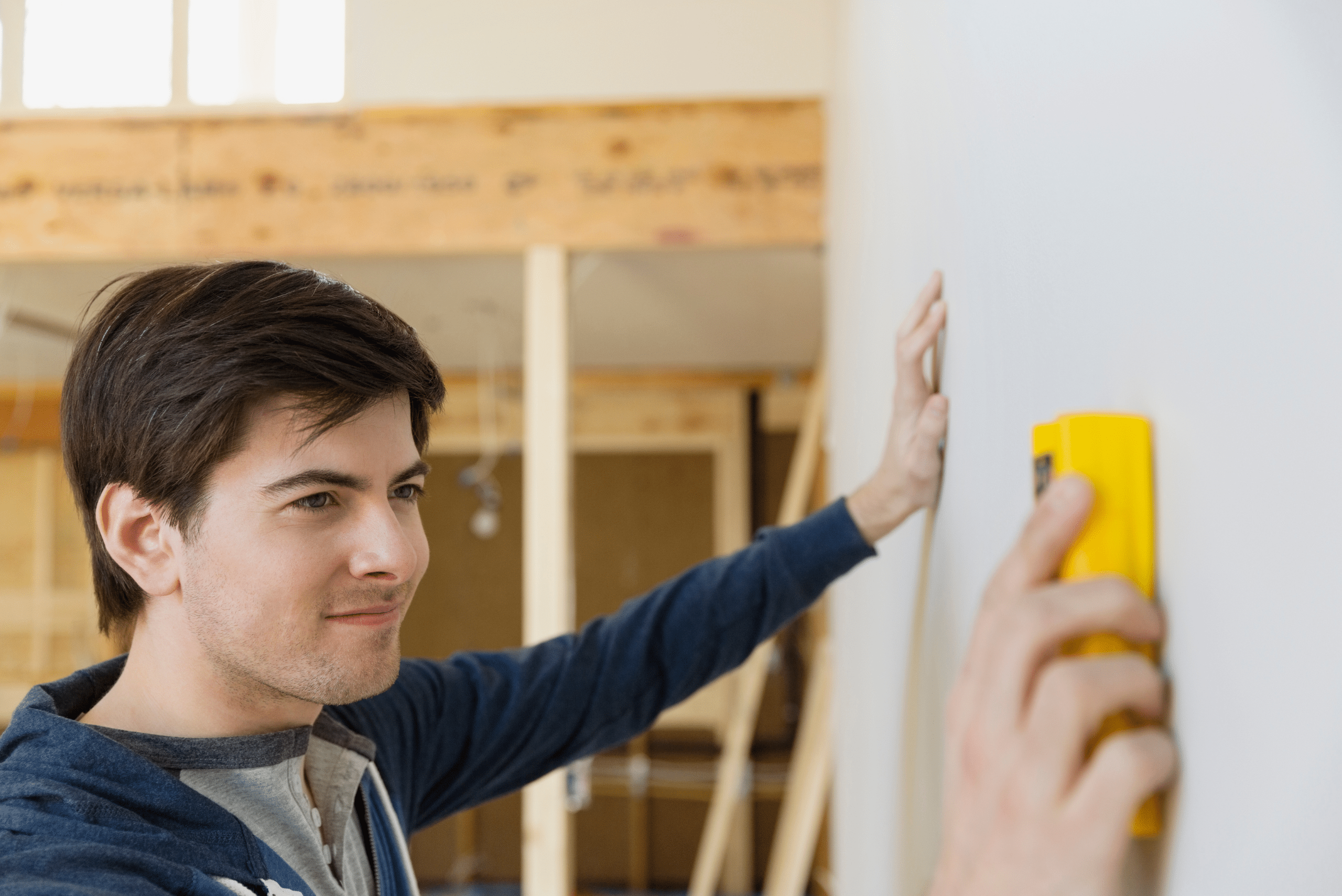 Man using a yellow stud finder on wall while placing other hand on wall.