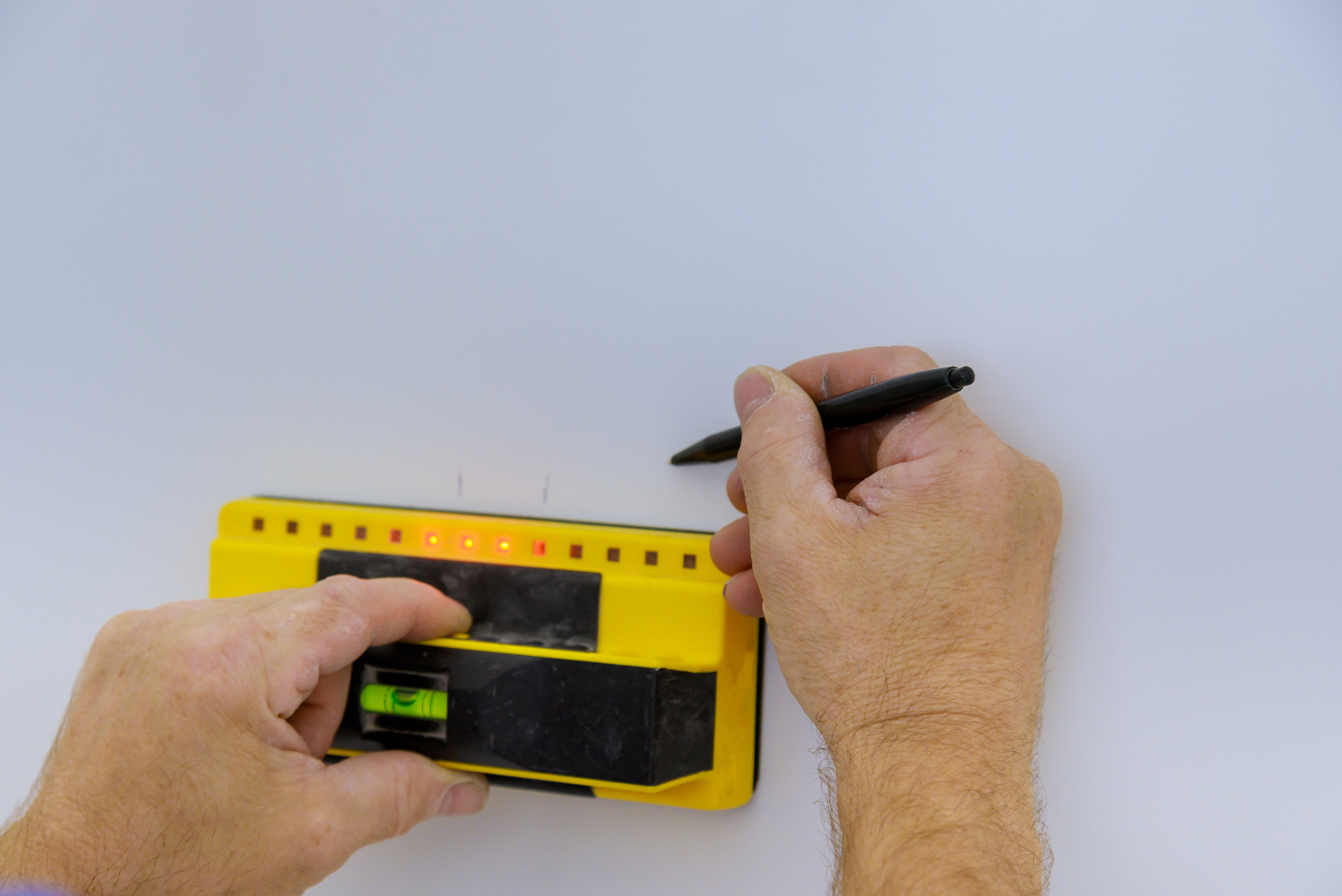 Hands of a person using a stud finder and pen to mark the wall for studs.