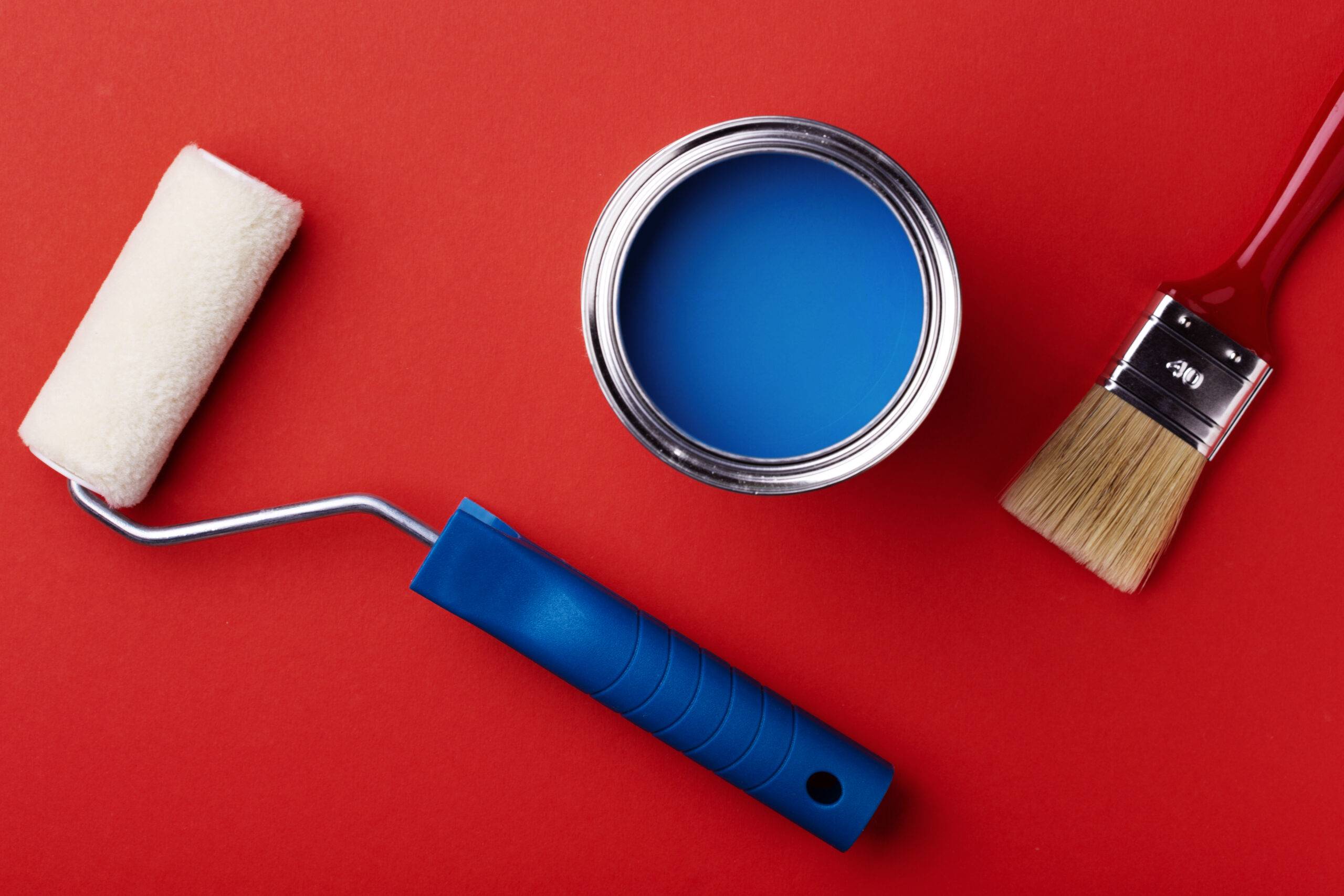 Can of blue paint with brush and paint roller on red background.