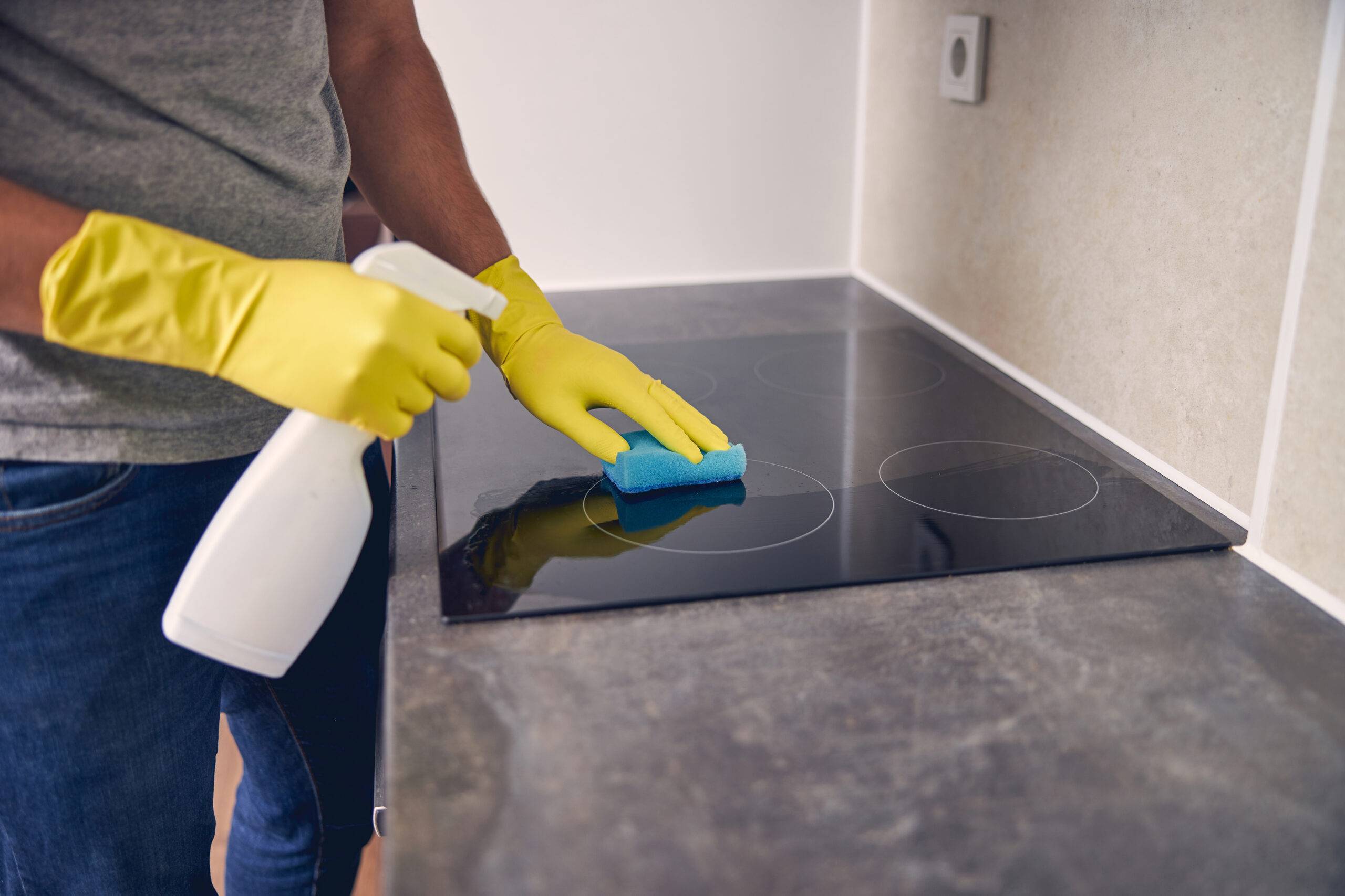 Man wearing yellow gloves to clean glass stove top using spray and sponge.