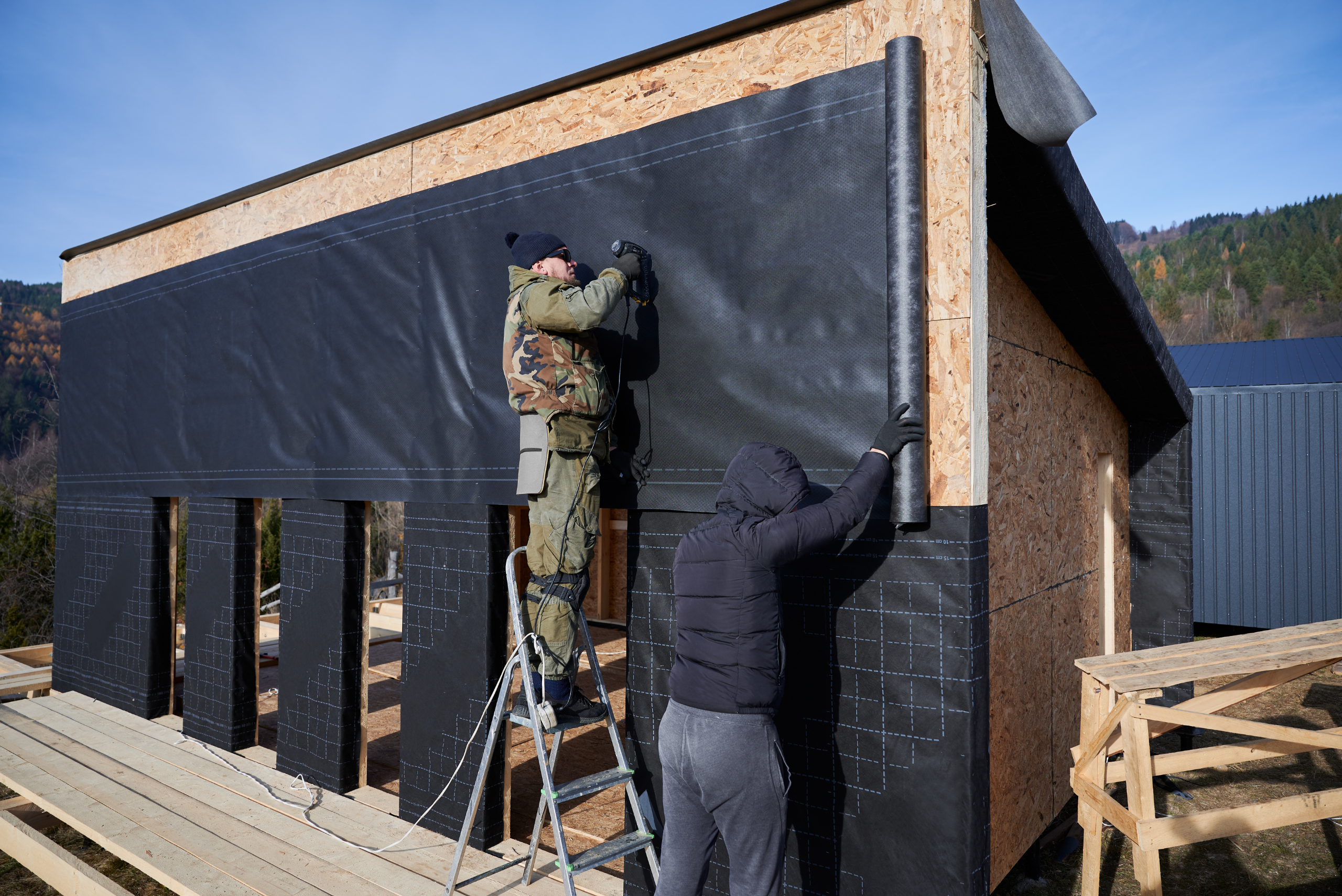 Male builders installing waterproof membrane on the wall of future cottage. Men workers building wooden frame house.
