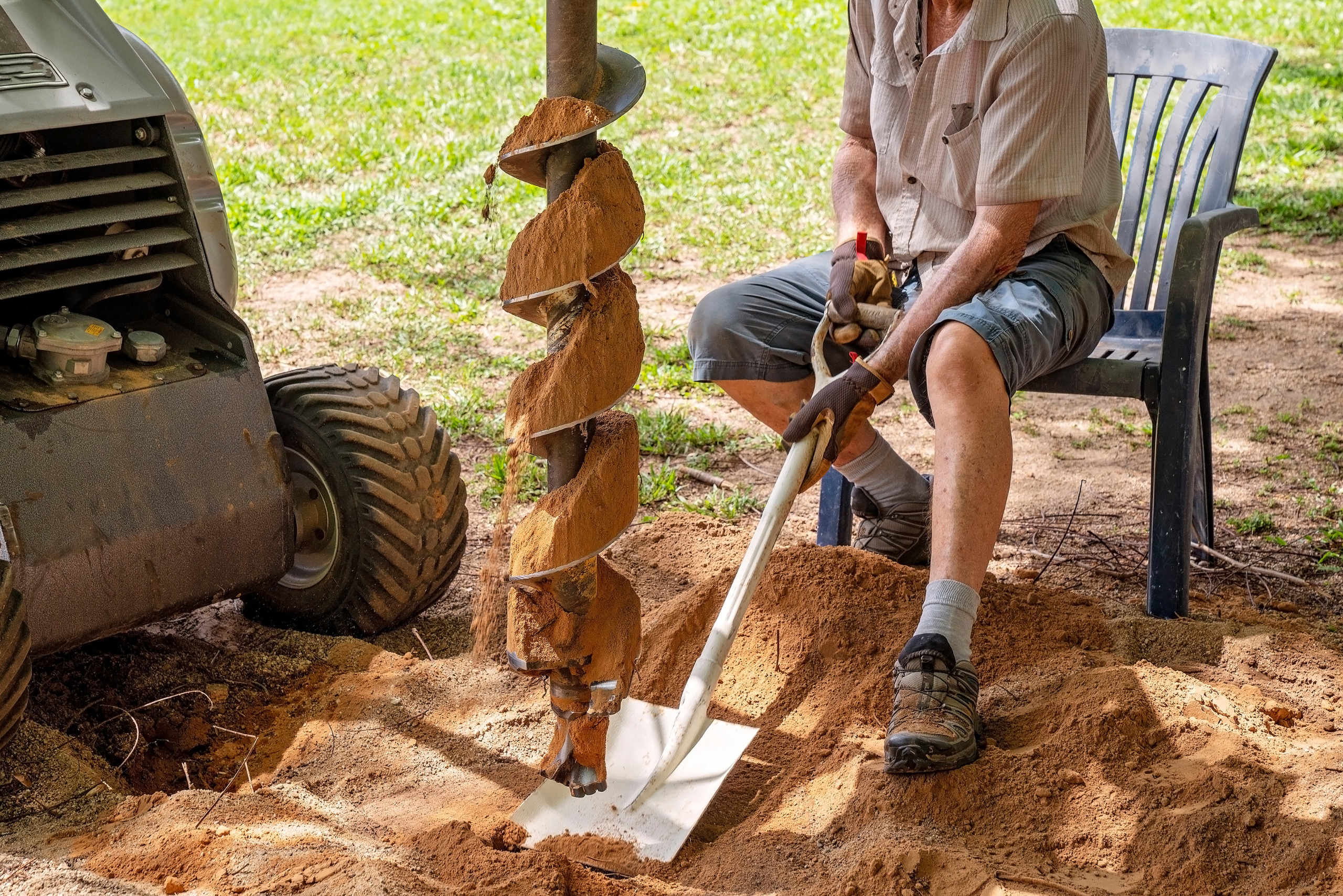 Person sitting on a plastic chair holding a shovel under a mechanical post hole digger bit.