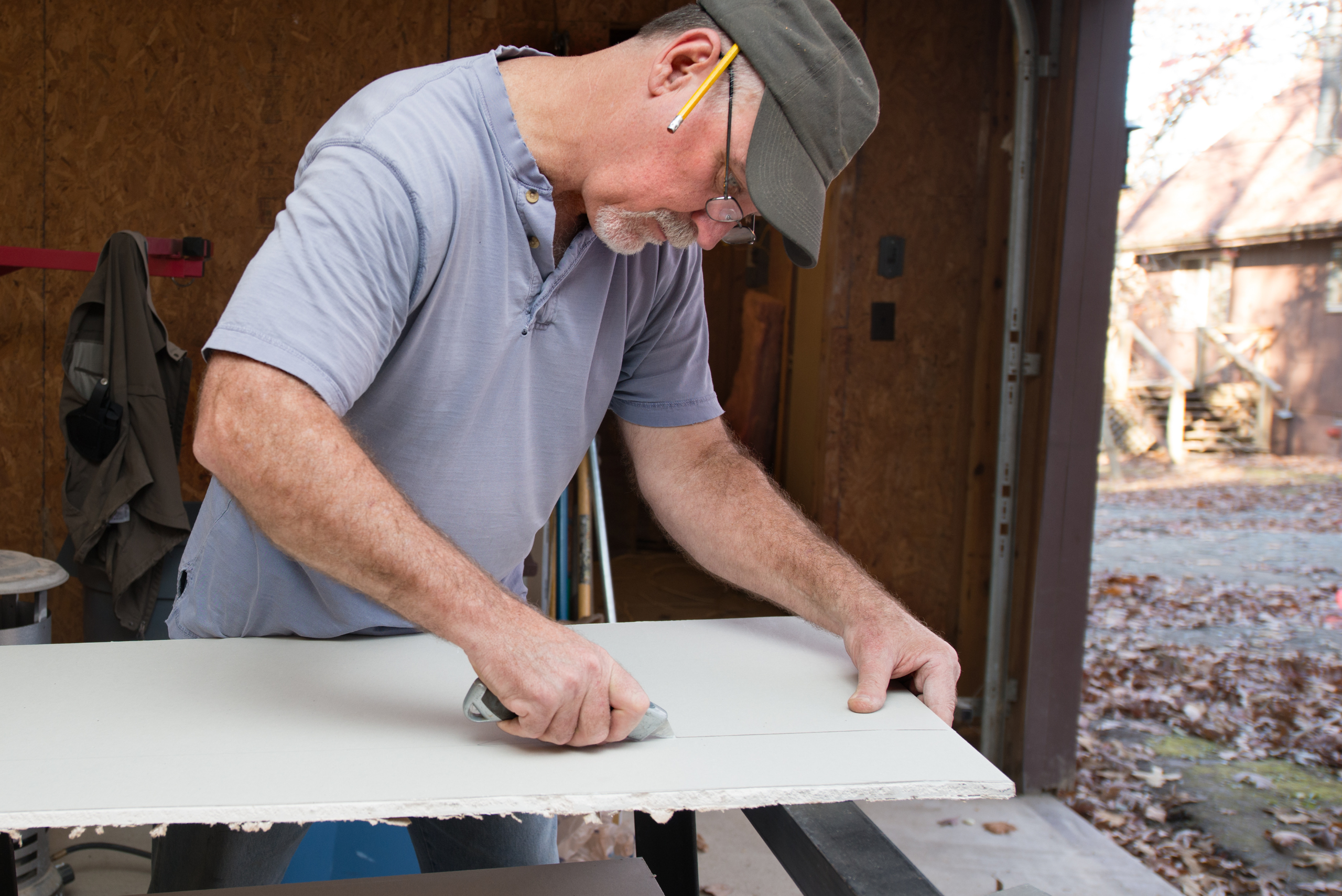 Person wearing a hat is holding and cutting a drywall board.