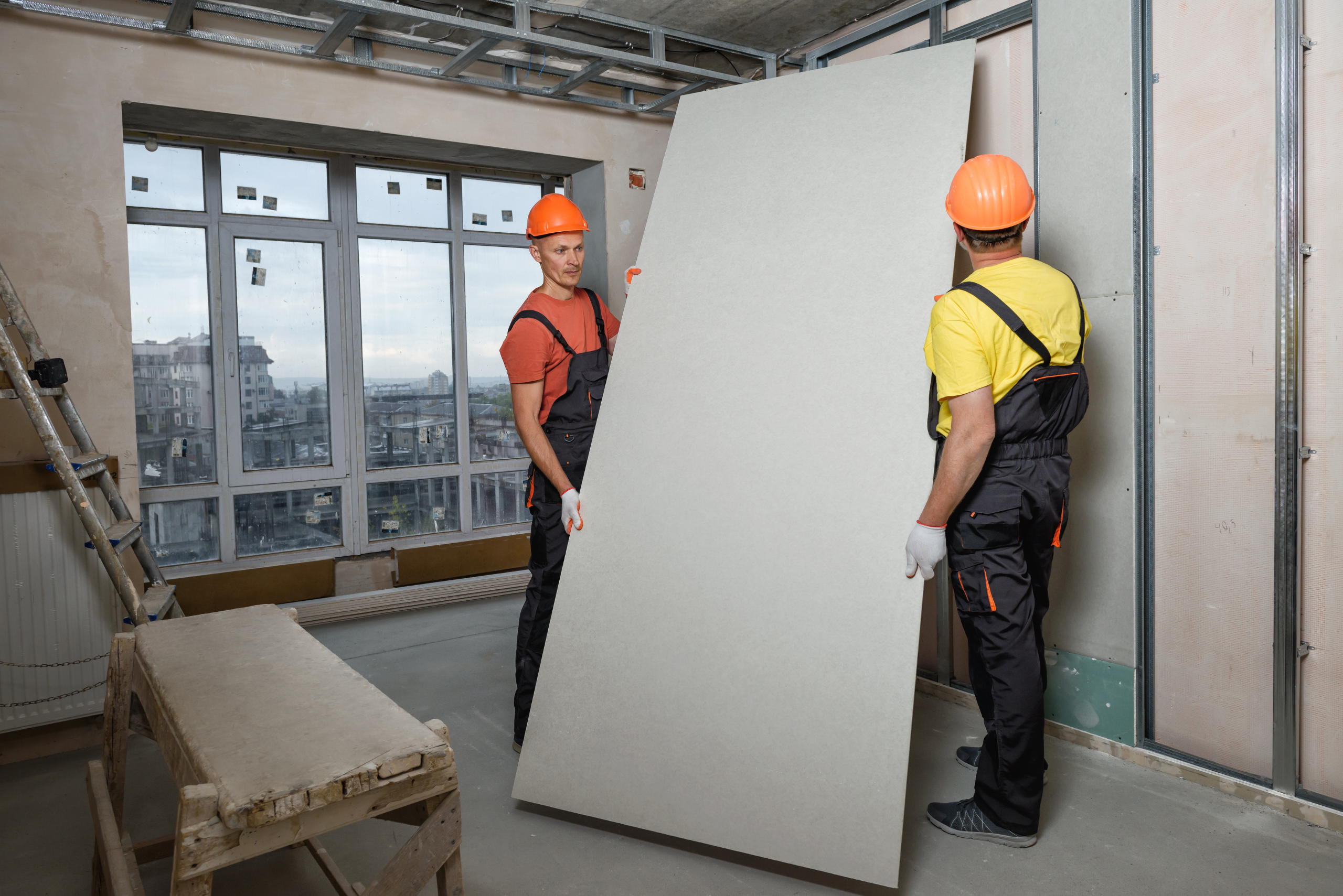 Two people wearing safety hard hats carrying a large board of drywall.