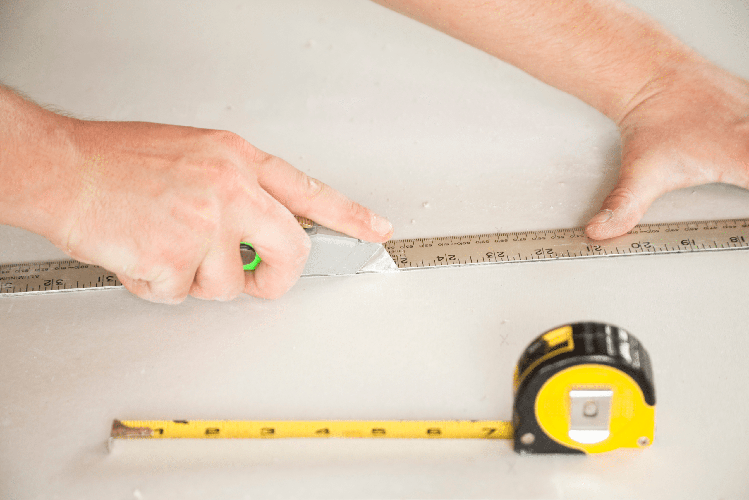 Closeup of hands, ruler, and tape measure. The right hand is holding a knife that is guided against the ruler supported by the left hand.