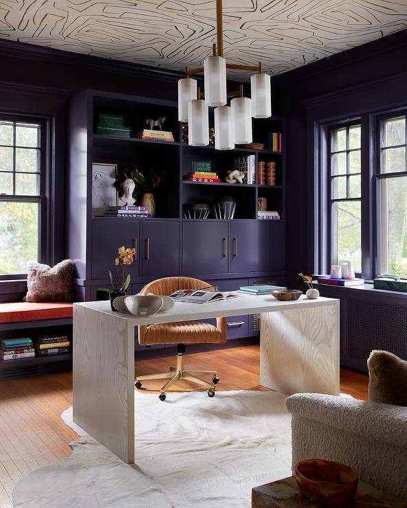 A gold and frosted glass chandelier hangs from a white and black wallpapered ceiling in a purple home office boasting a white oak waterfall desk matched with a white cowhide rug. The chair is placed in front of a purple built in cabinets topped with styled purple shelves. A purple window seat is topped with an orange cushion and a gray fur pillow. Purple walls are lined with purple crown moldings.