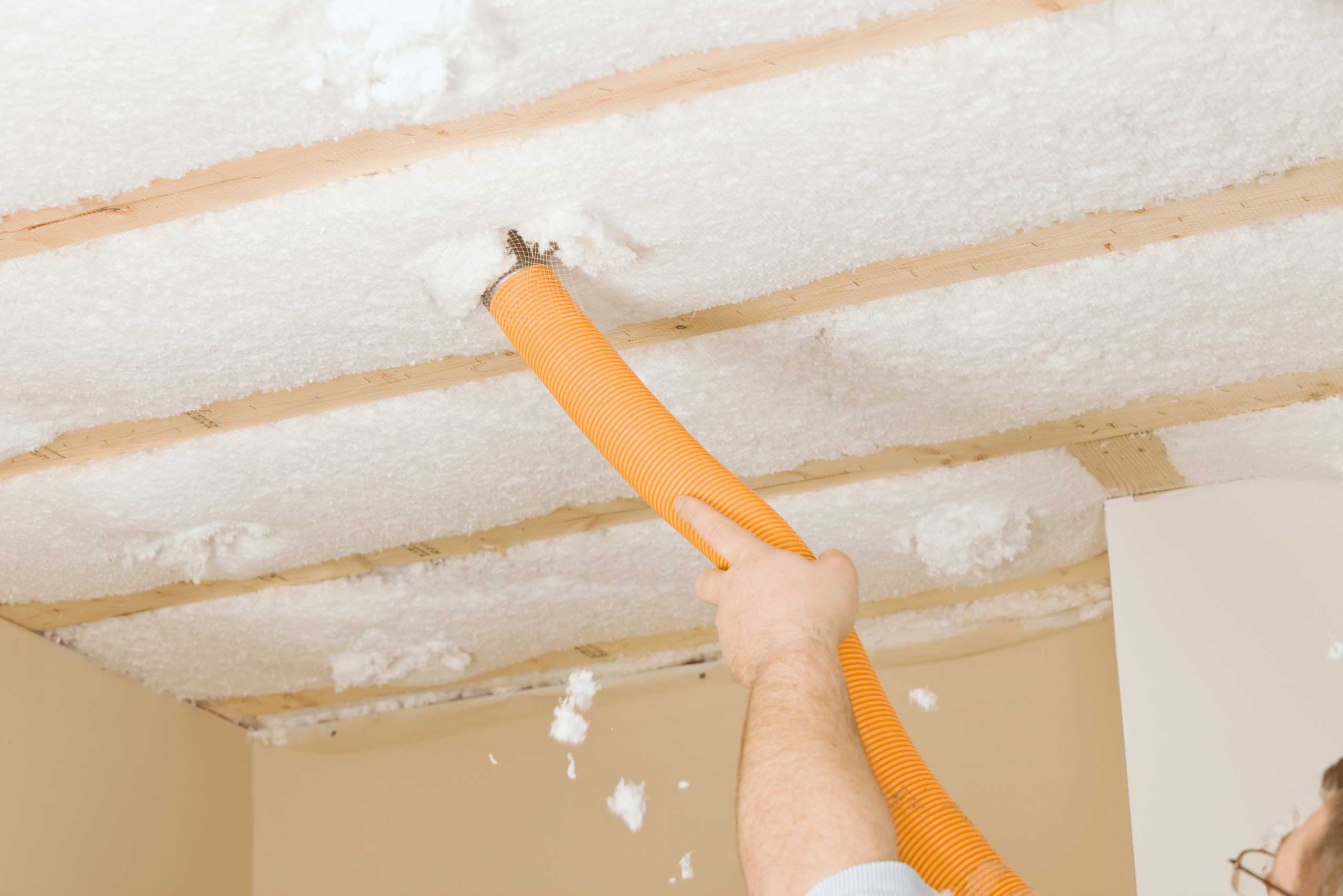 A closeup of someone's hand holding an orange tube that is pushing out insulation into the basement ceiling.