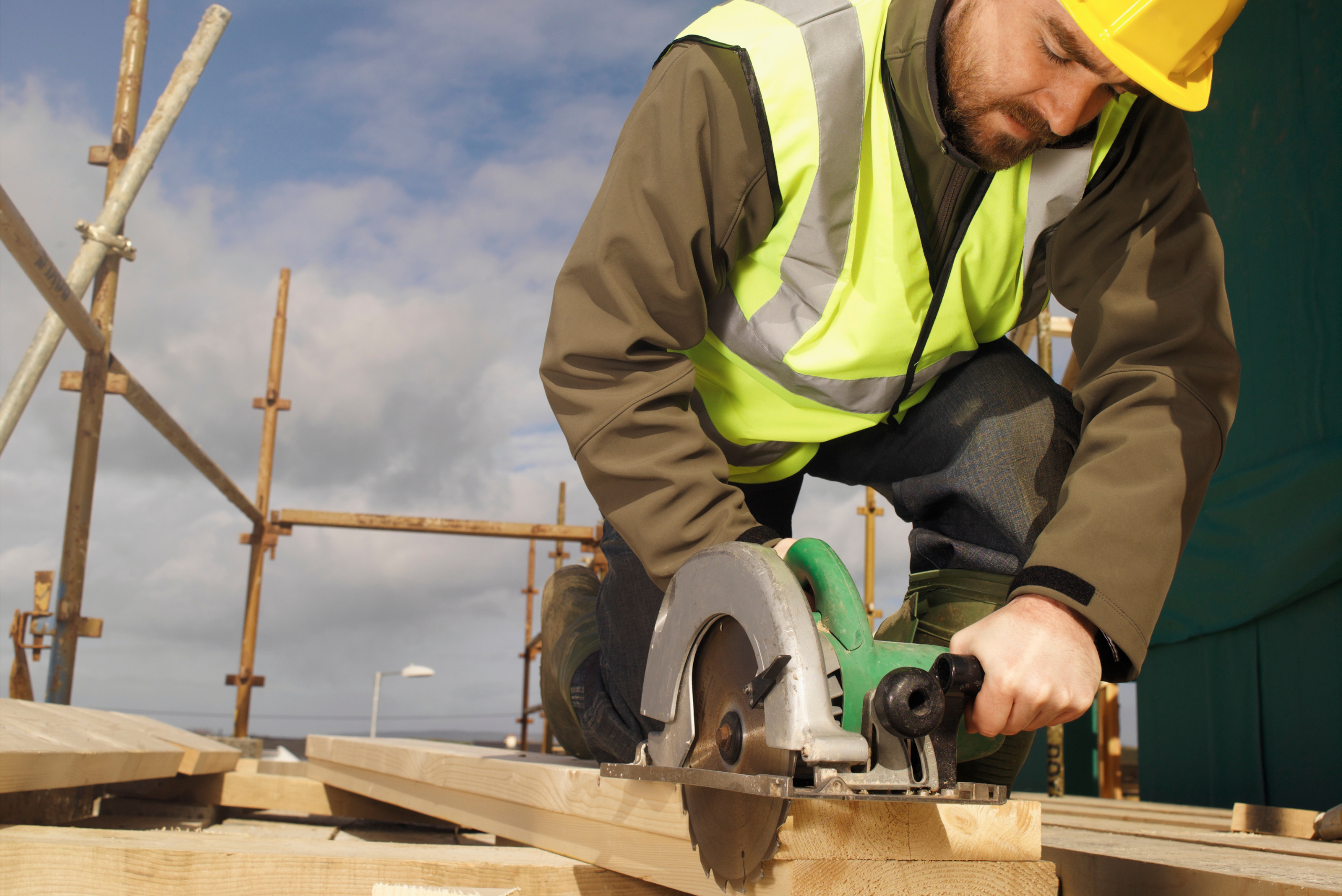 A person wearing safety hat and vest while holding a circular saw with both hands to make a cut in wooden board.