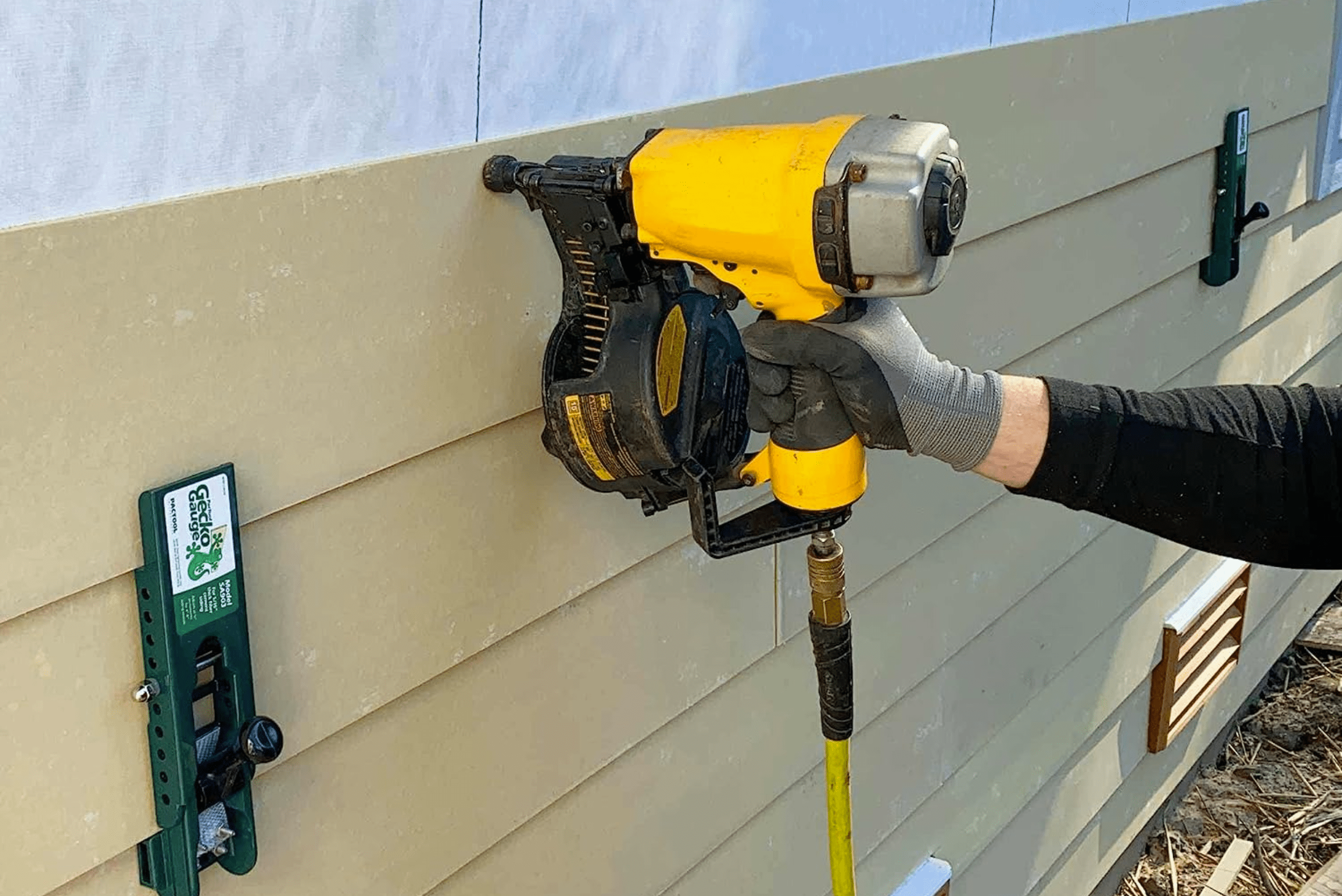 A closeup of a nail gun securing hardie boards to the side of a house.
