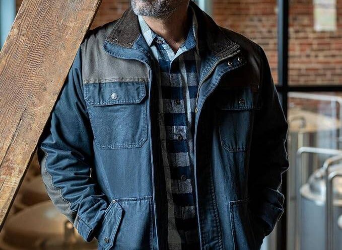 A man leaning on wooden beam while wearing a chore jacket.