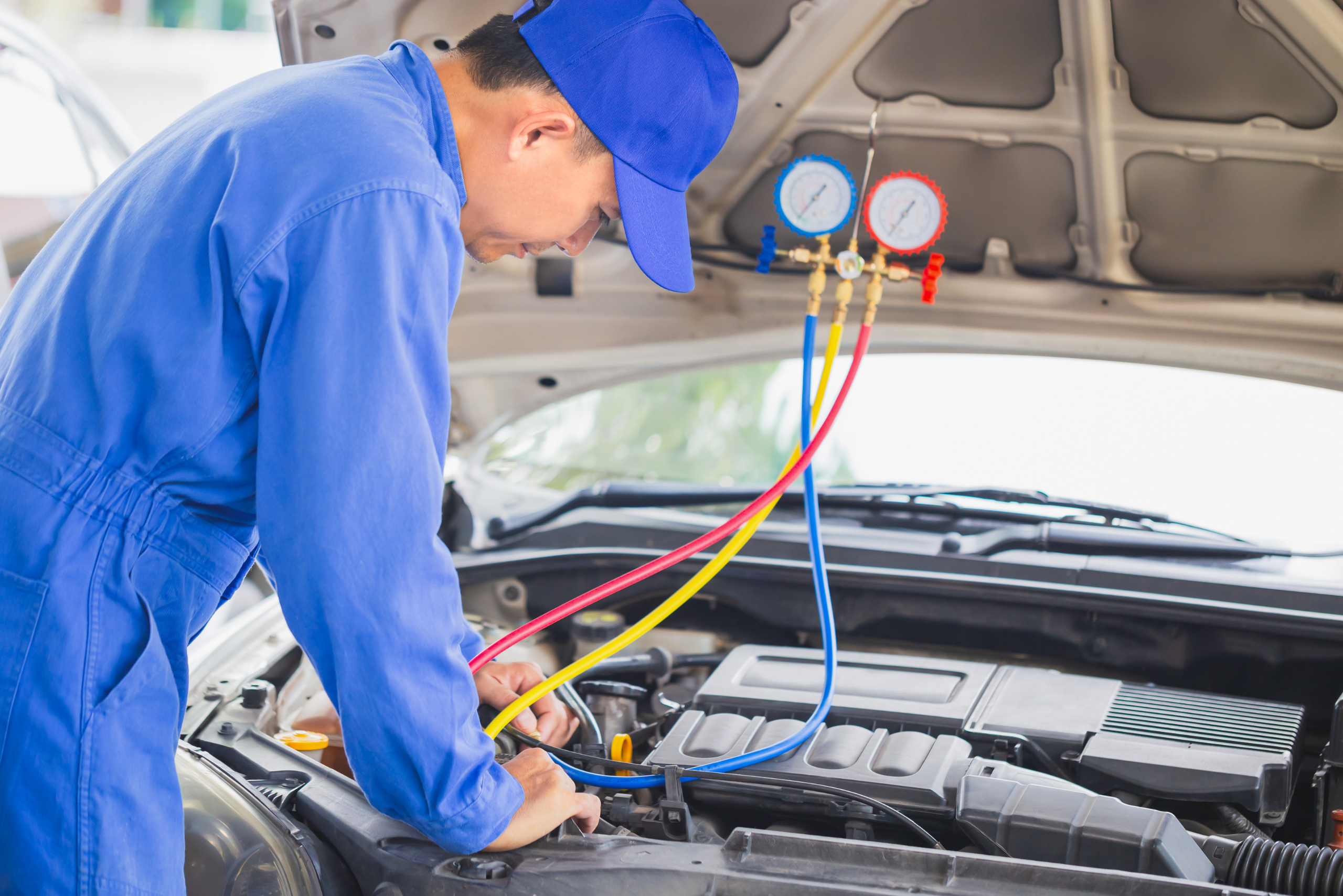 A person in all blue using an AC recharging tool under the hood of a car.