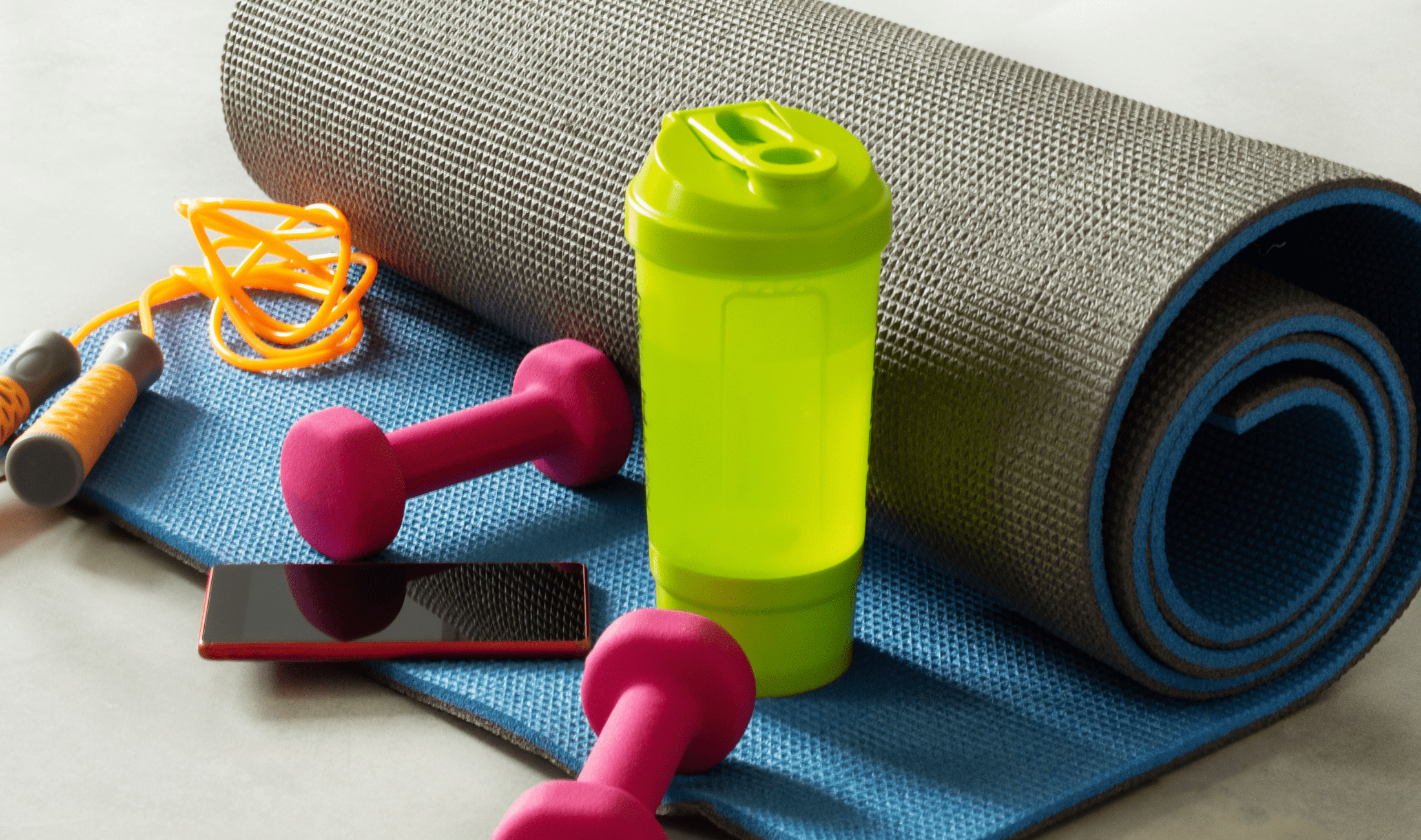 A staged photo of a rolled up yoga matt, two small hand weights, a shaker bottle, a jump rope, and a smart phone.