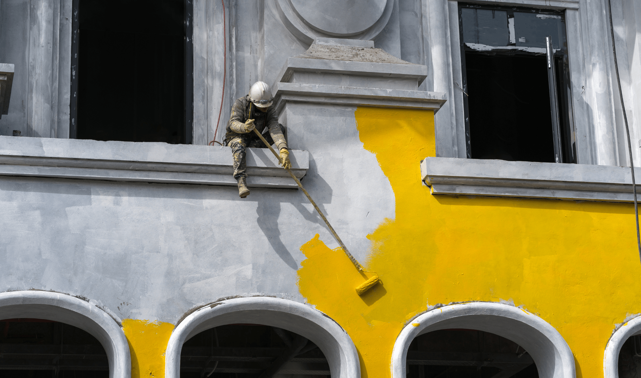 A man sitting on a building ledge using a long paint roller to paint the exterior of a building yellow.