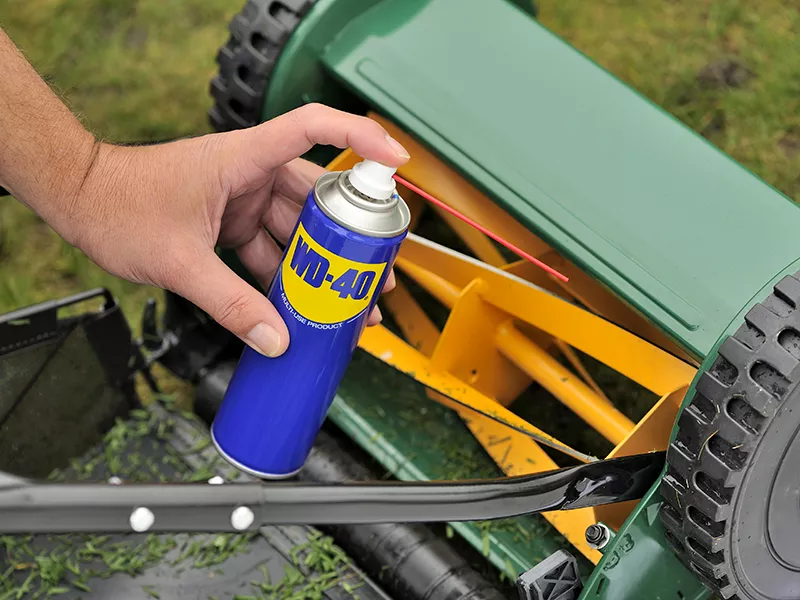 Someone holding a can of WD-40 above lawnmower blades.
