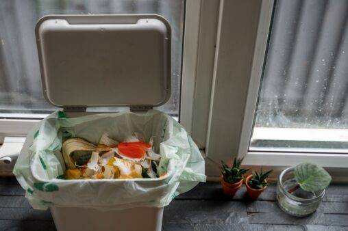 An open garbage bing with some food garbage at the top next to a window.