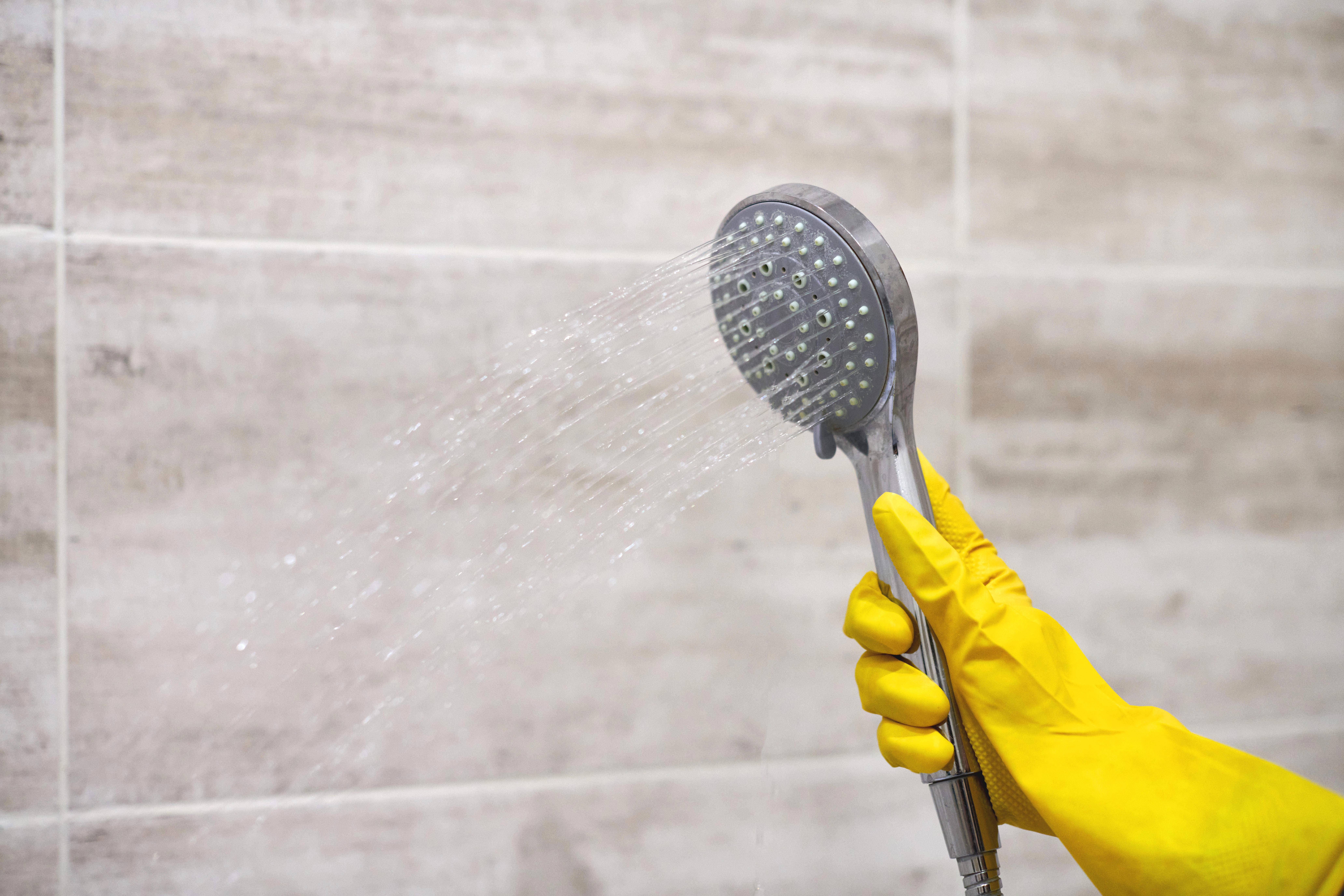 Female hand in protective yellow rubber glove holding shower head with pouring water in domestic bathroom, copy space, motion blur.