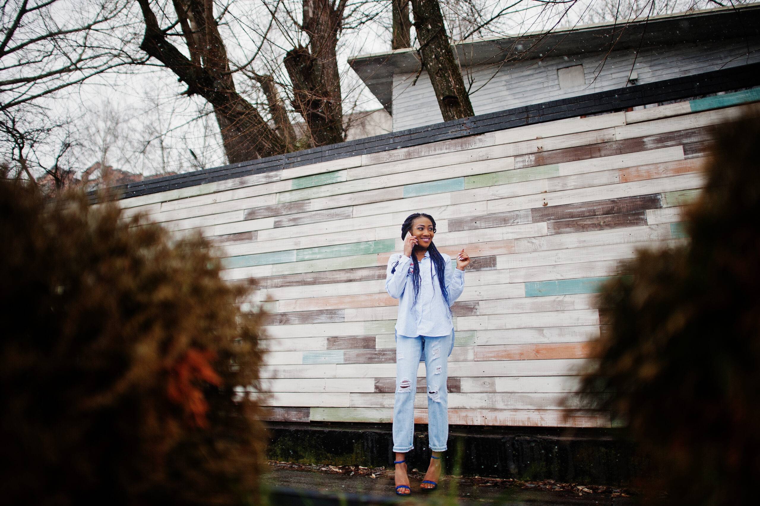 Stylish african american girl with dreads holding mobile phone at hand, outdoor against wooden wall.
