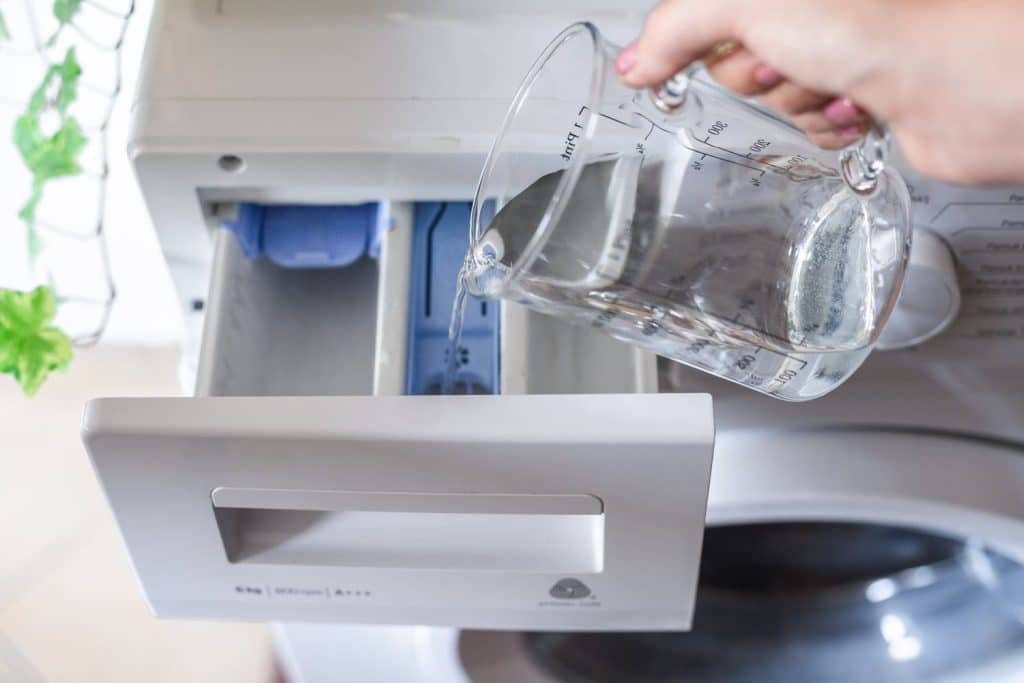 Someone pouring a container of vinegar into the detergent slot of a washing machine.