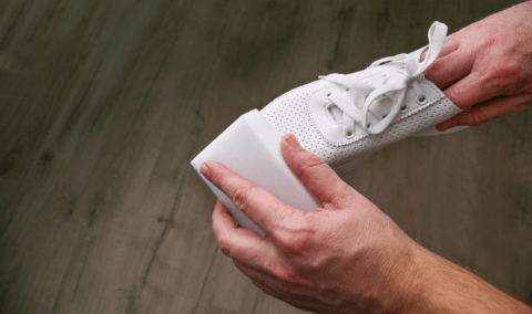 A pair of hands, one holding a shoe, the other cleaning it with a melamine sponge.